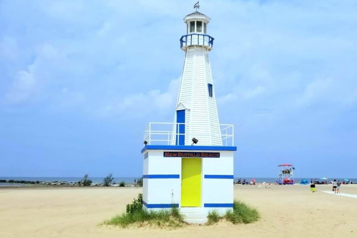White and blue decorative lighthouse structure on New Buffalo City Beach with sandy shoreline and beachgoers