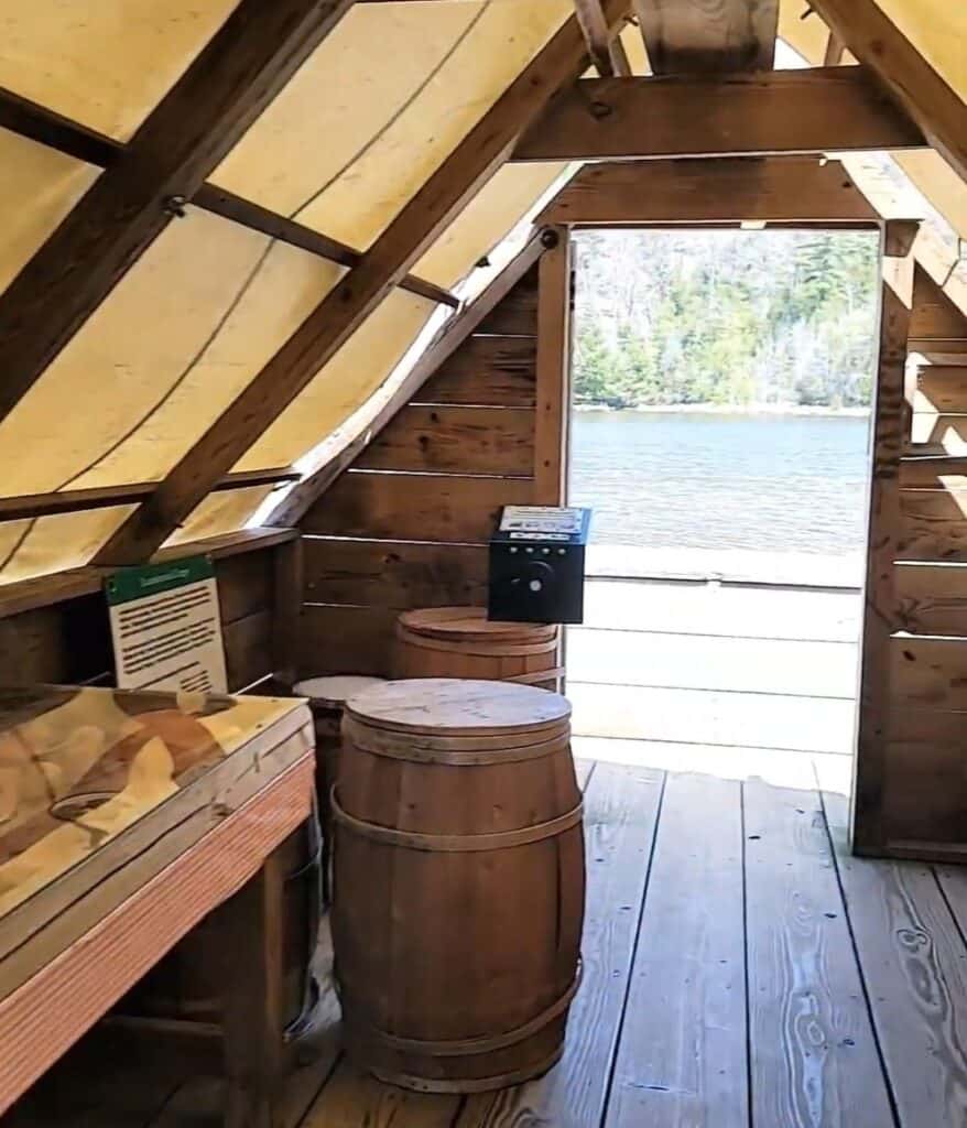 Interior of the wanigan floating cook shack at Lumberman's Monument showing wooden barrels, interpretive display, and AuSable River through the doorway