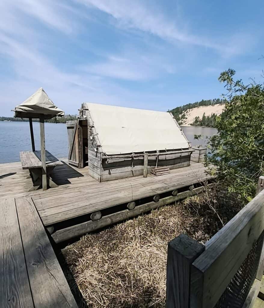 Wanigan floating cook shack replica on a wooden platform at the base of the 260 steps at Lumberman's Monument, with the AuSable River and sand dune behind