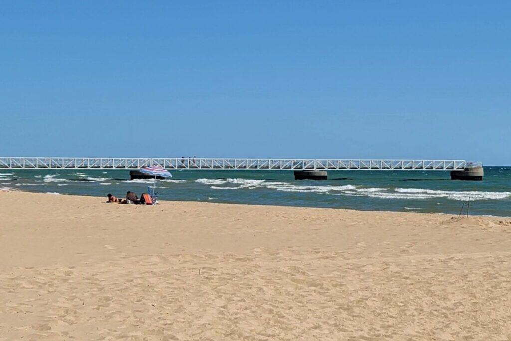 Long white pier extending into Lake Huron at Oscoda Beach Park with sandy beach and umbrella in the foreground